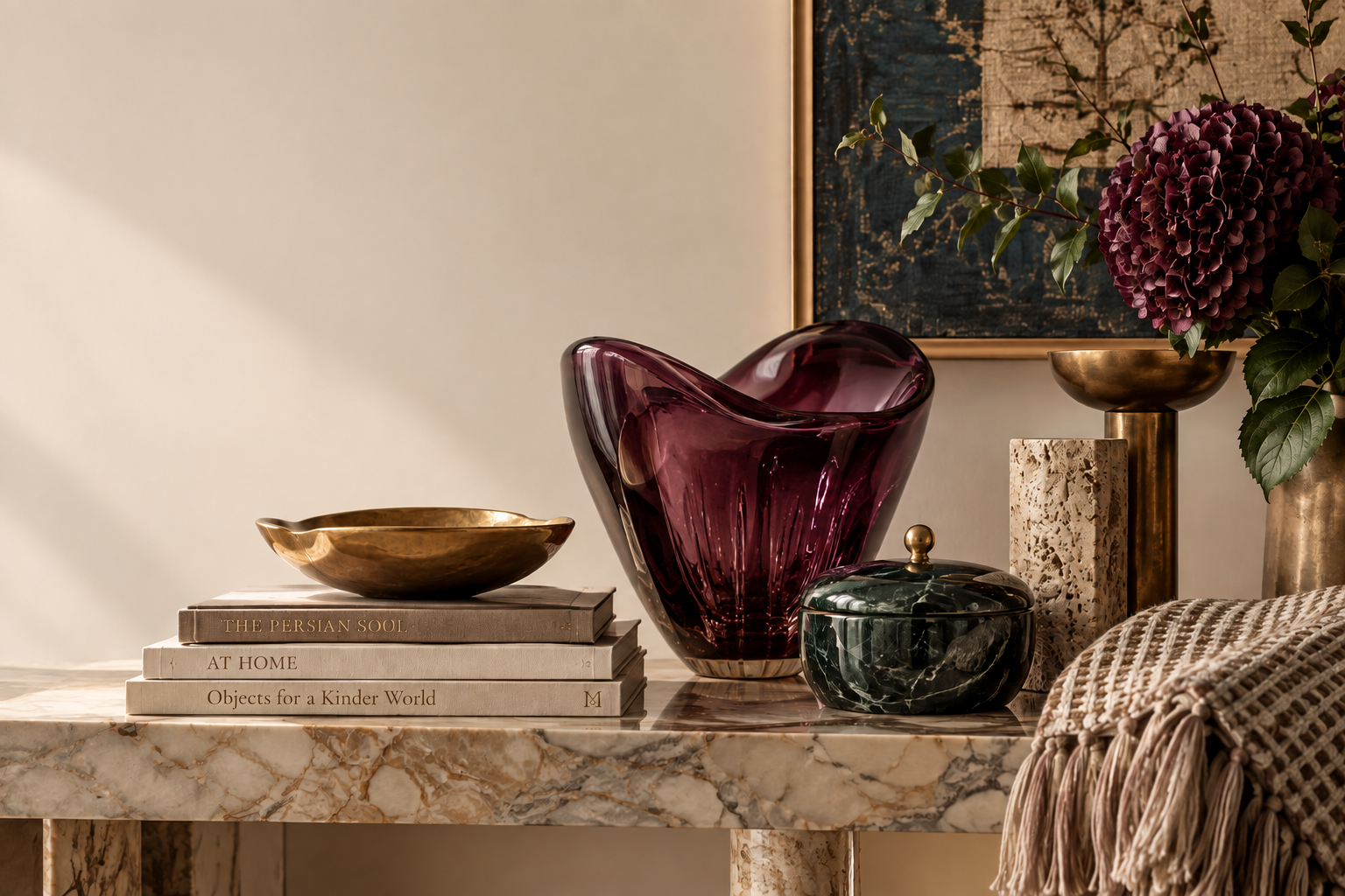 A veined marble console styled with a deep purple glass vase, a dark green marble jar, brass bowls, maroon hydrangeas and a stack of books